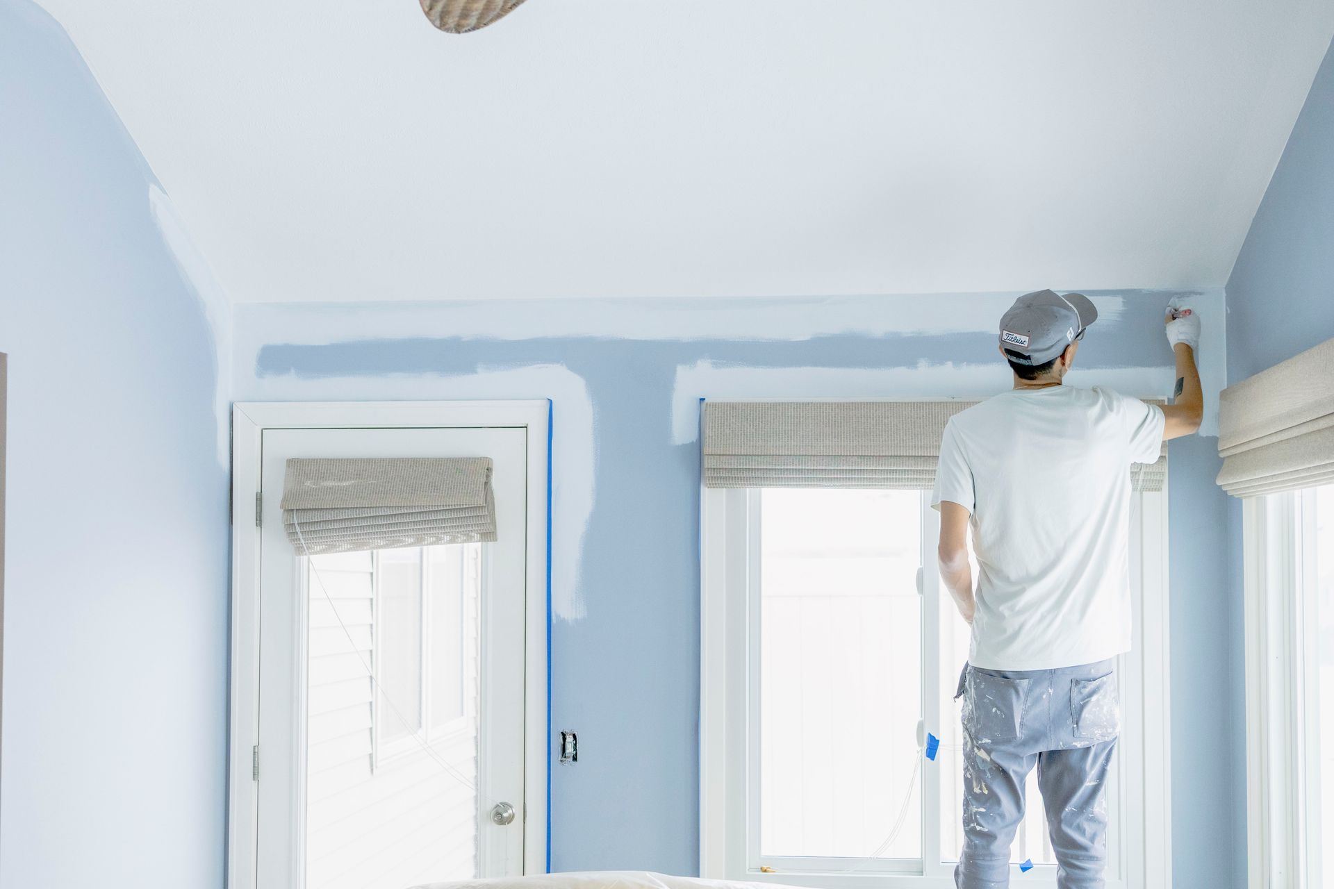 A person wearing a white t-shirt and grey cap paints the upper wall of a room with white paint, covering light blue walls.