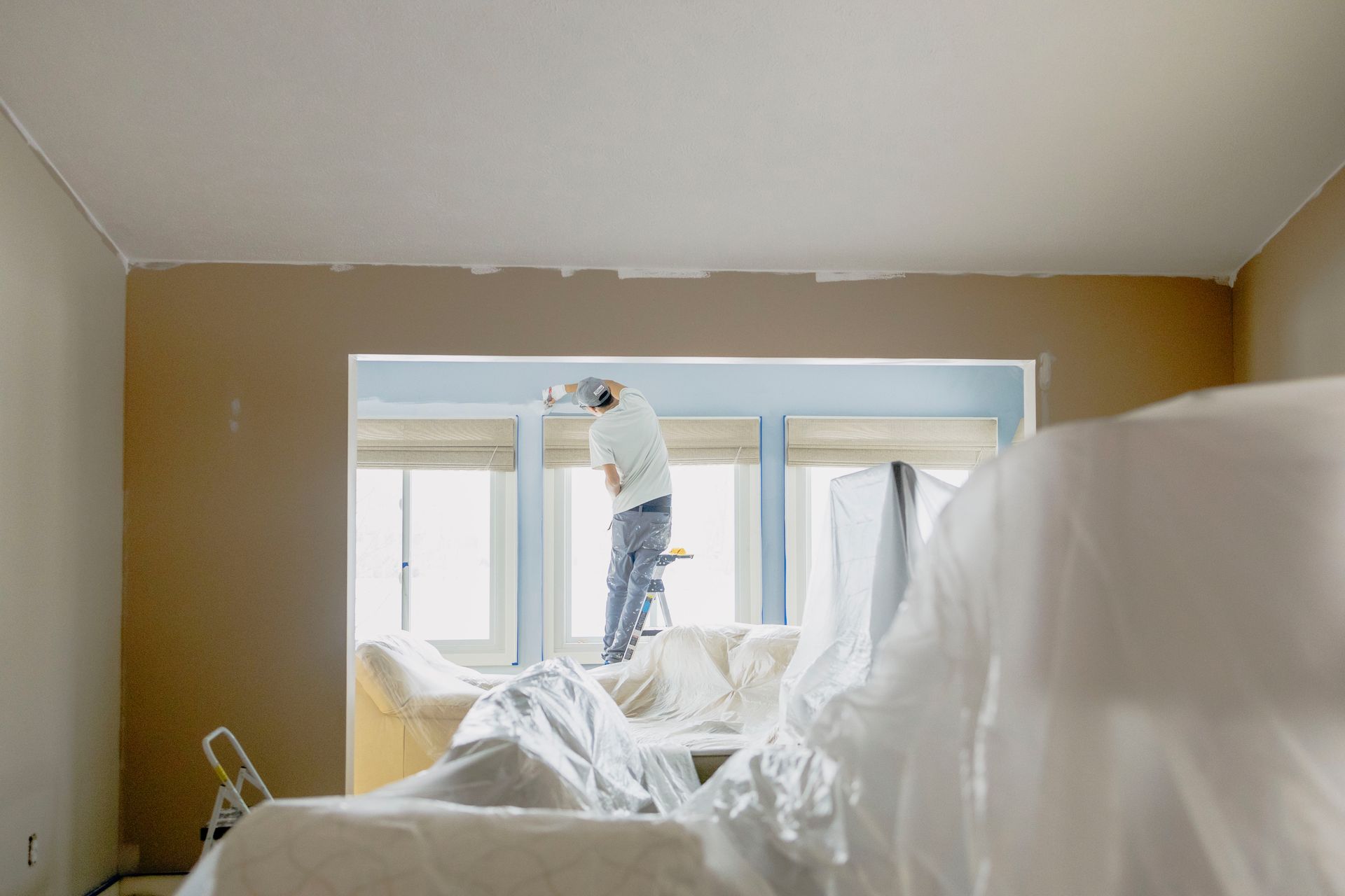 A person stands on a small ladder, painting the trim around a large window in a room undergoing renovations.