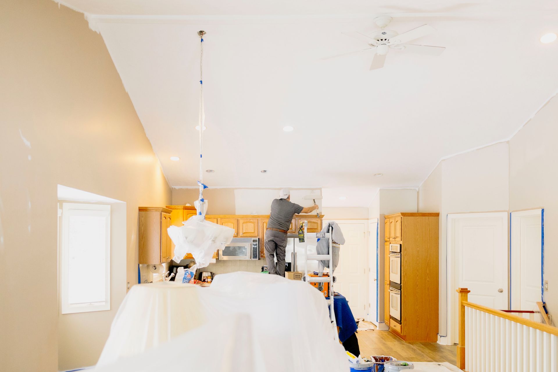 A person works on a kitchen ceiling renovation, with plastic sheeting covering the area below in a brightly lit home.