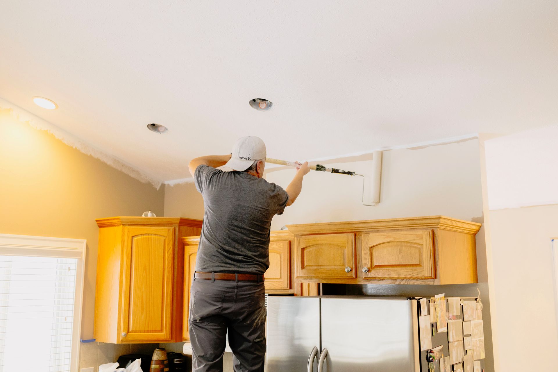 A person in a gray shirt stands on a ladder using a roller to paint the upper wall of a kitchen with wood cabinets.