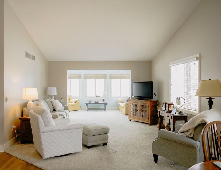 A spacious living room with cream walls, beige carpet, two armchairs, a sofa, and a television on a wooden stand.