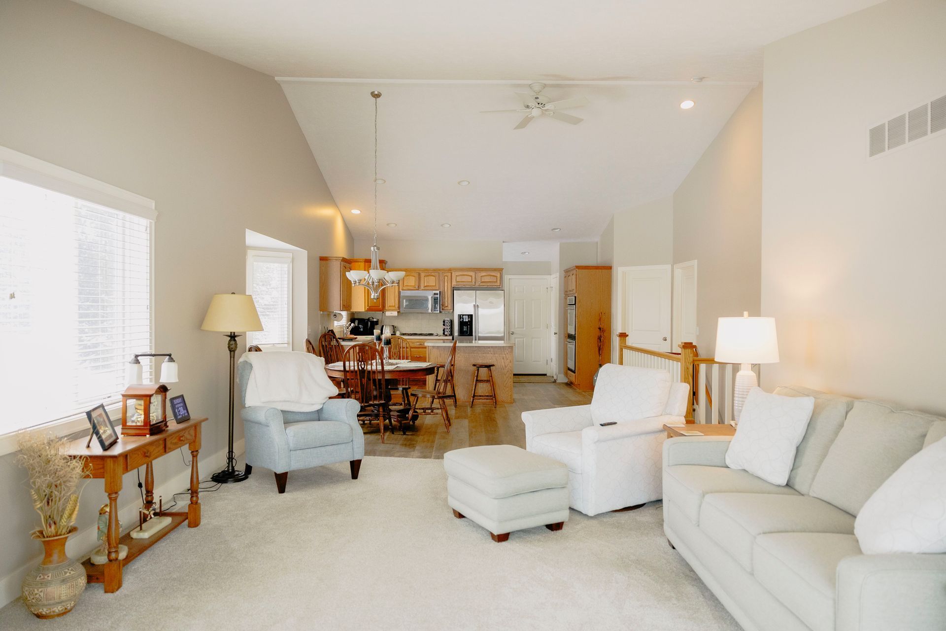 A living room with light walls and carpet, featuring a white sofa, armchair, ottoman, and an open kitchen in the background.