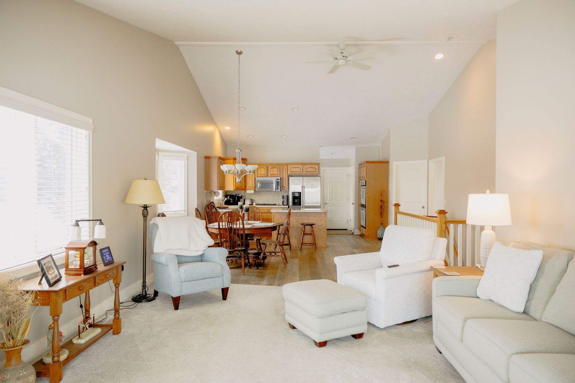 A bright, neutral-toned living room opening into a dining area and kitchen with light wooden cabinets and vaulted ceilings.
