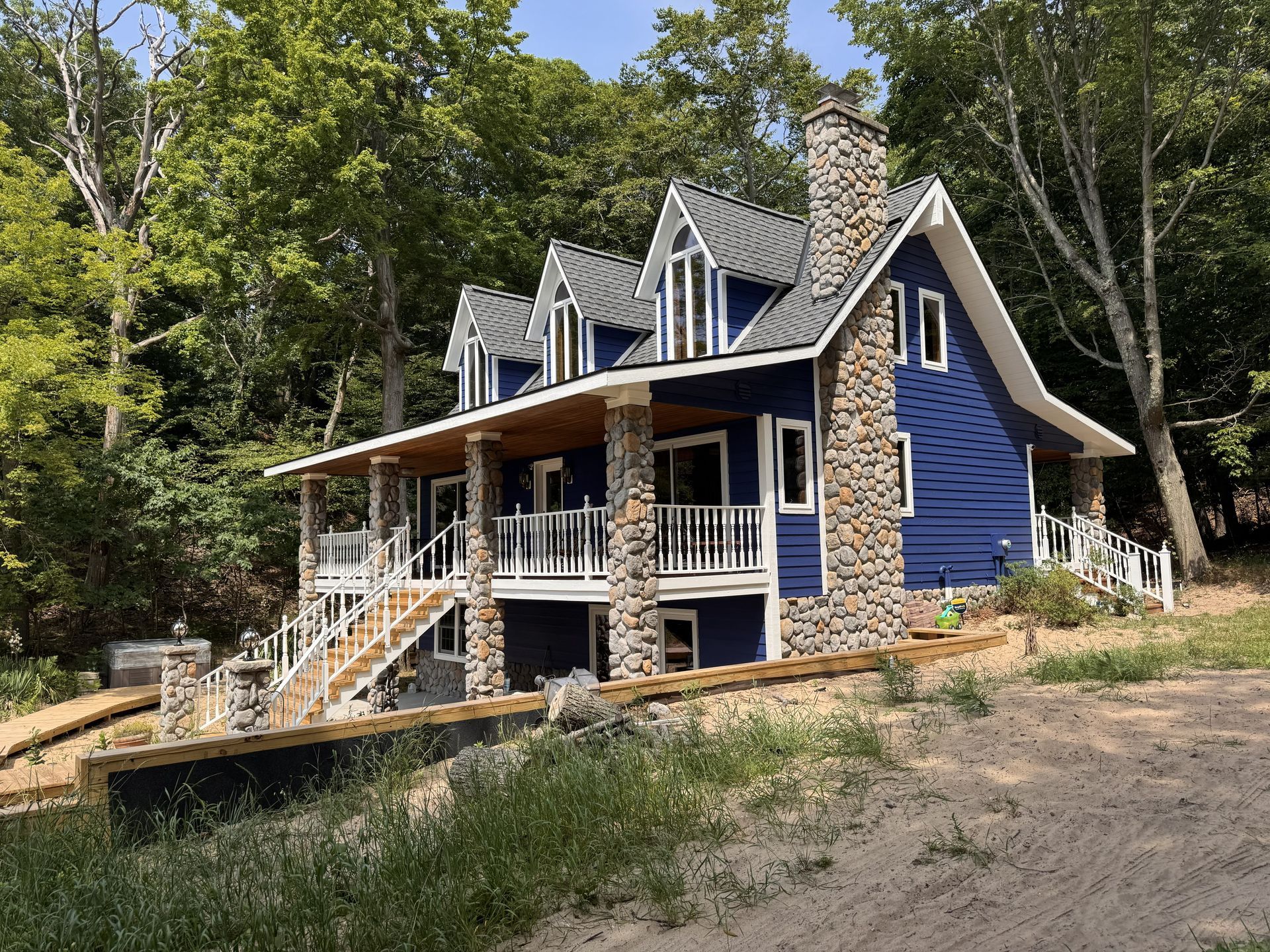 A two-story blue house with a stone chimney, wrap-around deck, and three roof dormers nestled among lush green trees.