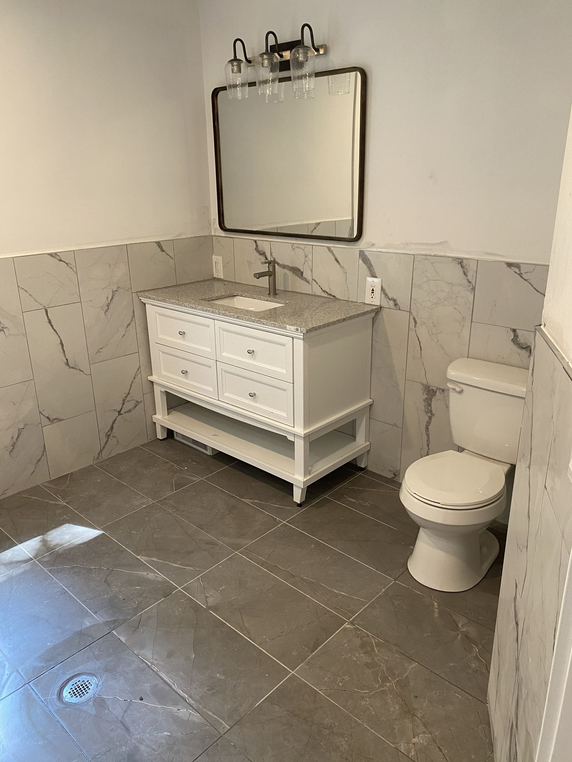 A bathroom with grey tiled floors, a white vanity with a mirror and lighting, and a white toilet against marble walls.