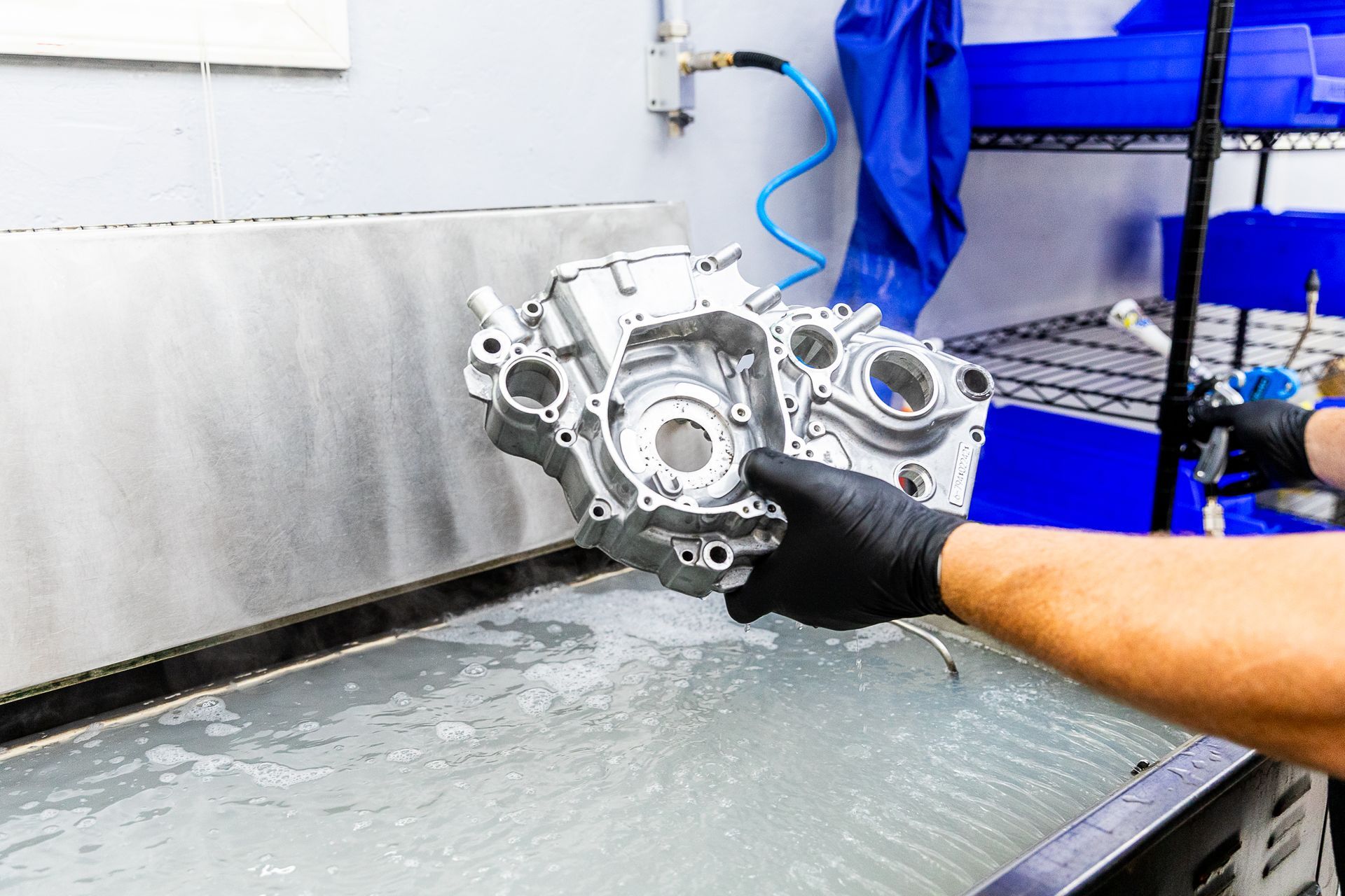 Gloved hand holding a metal engine part over a workshop cleaning tank