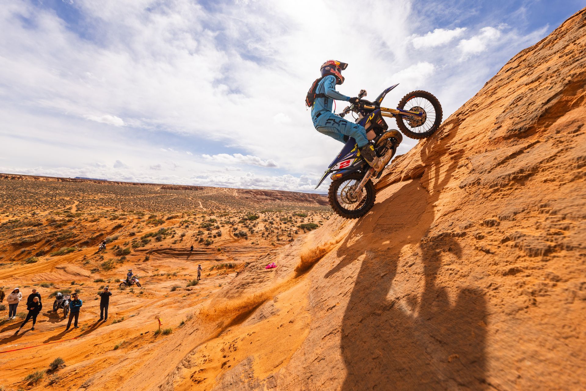 Mountain biker climbing a steep orange desert rock face with spectators below under a blue sky