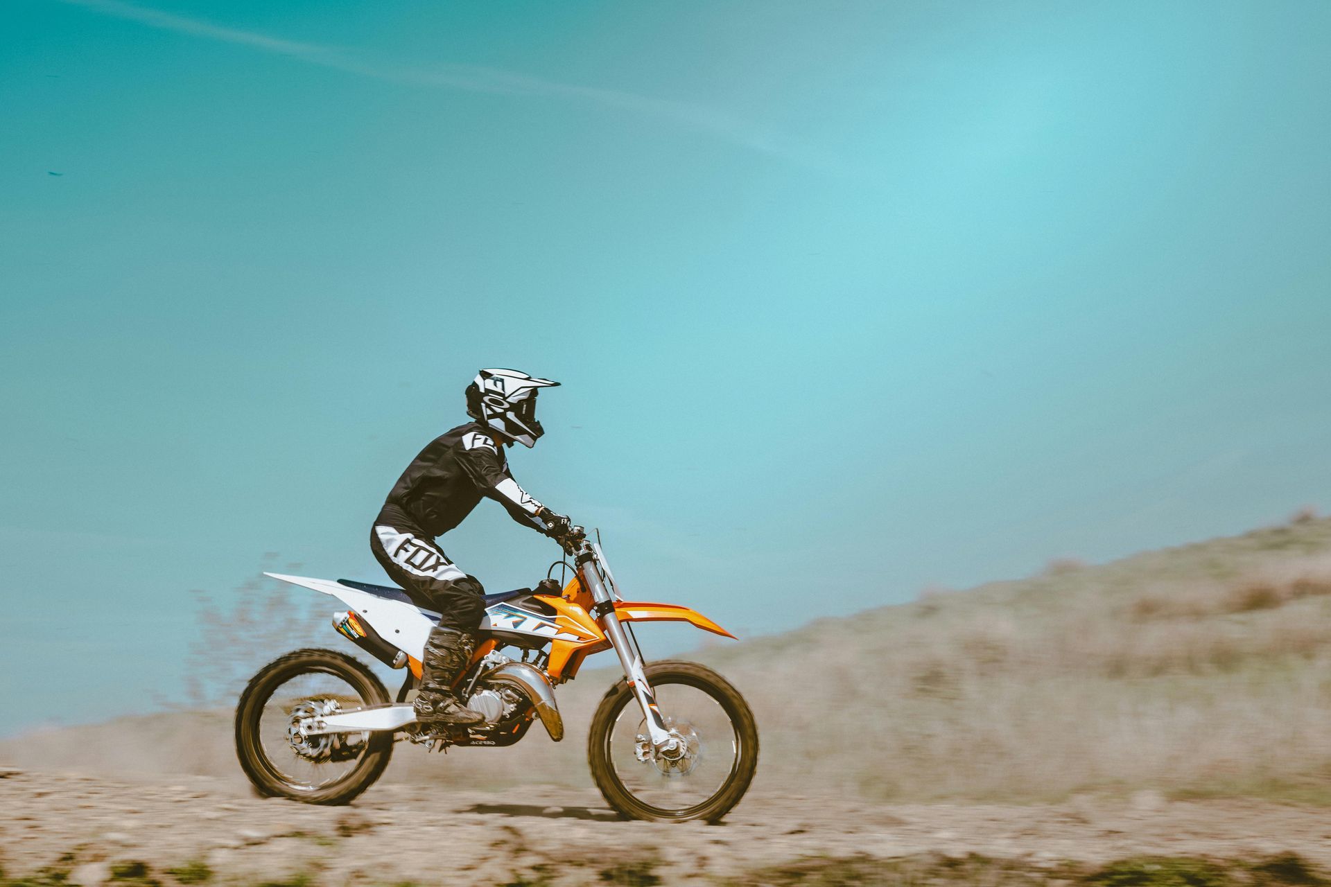 Motocross rider on an orange dirt bike riding across a dusty desert track under a blue sky