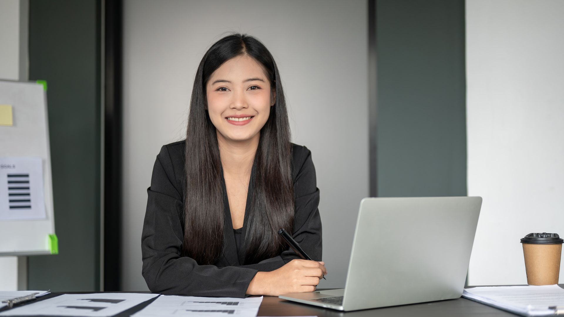 Woman at a desk with a laptop and coffee cup, smiling in a modern office.