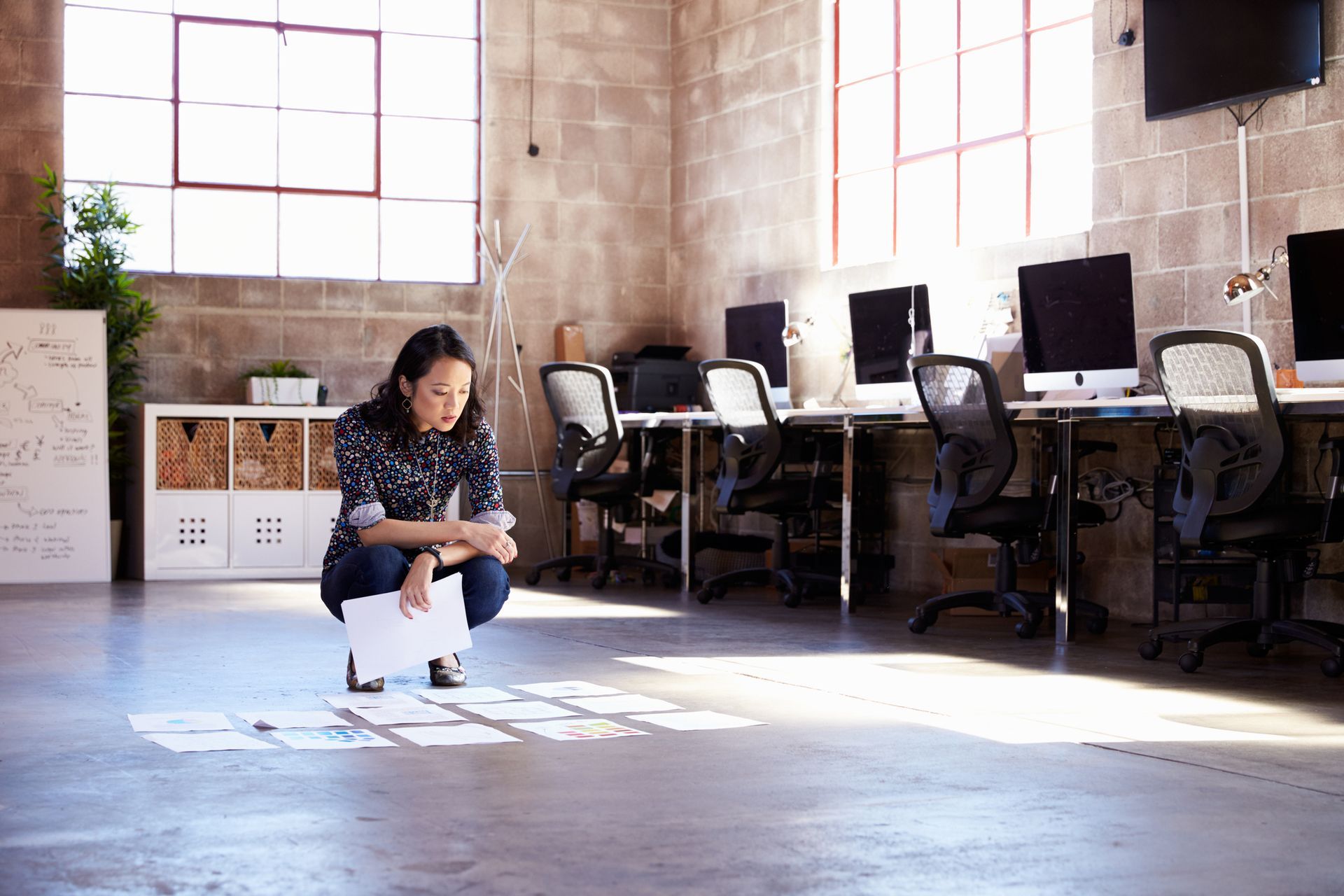 A woman is squatting on the floor in an office looking at papers.