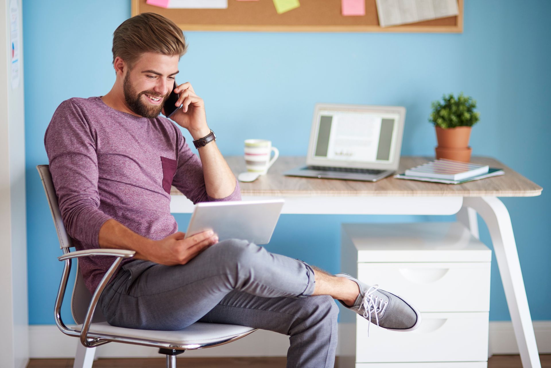 A man is sitting at a desk talking on a cell phone while using a laptop.