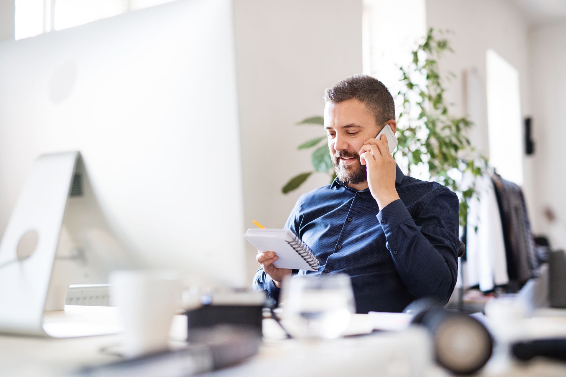 A man is sitting at a desk talking on a cell phone while holding a tablet.