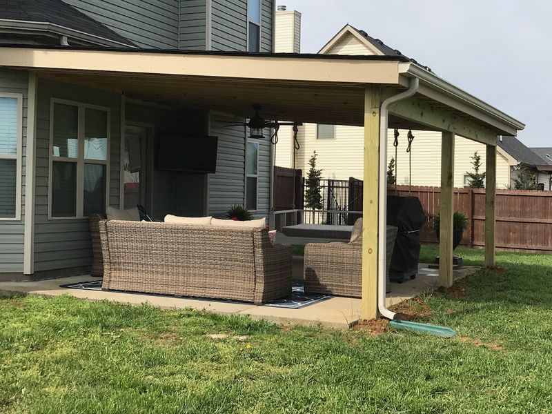 Covered backyard patio with wicker furniture, a grill, and green lawn beside a house