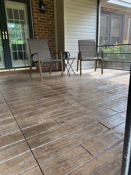 Covered wooden porch with two chairs and a small table beside a brick house and screened railing.