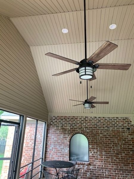 Covered patio with vaulted ceiling, two ceiling fans, brick wall, and a table by the window.