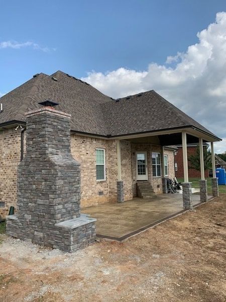 Brick house with a covered porch, stone chimney, and concrete patio under a cloudy sky
