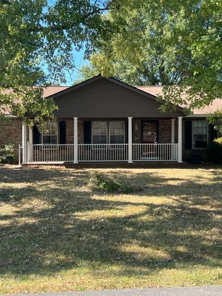 Single-story brick house with a covered front porch and white railing, framed by trees and a dry lawn.