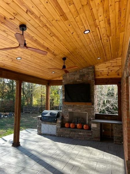 Covered patio with wood ceiling, stone fireplace, grill, and seating overlooking a wooded yard