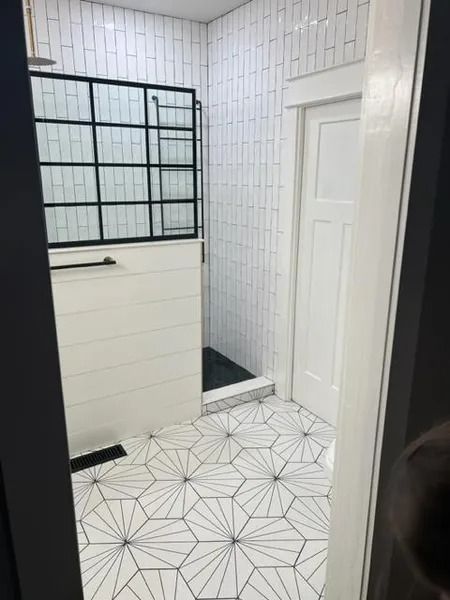 Small bathroom with white tiled walls and geometric floor, shower on left and white door on right.
