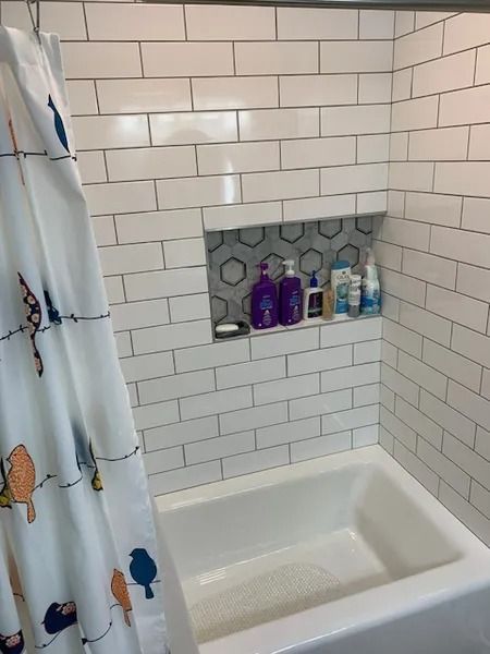 White-tiled bathroom shower tub with built-in shelf of toiletries and a patterned shower curtain