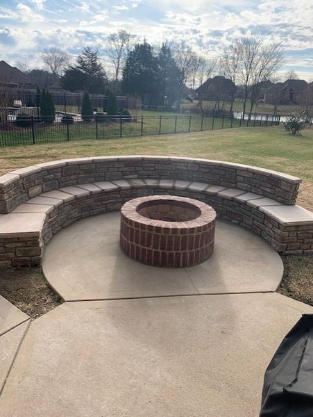 Brick fire pit surrounded by a curved stone bench in a park pavilion, with a grassy field behind it.