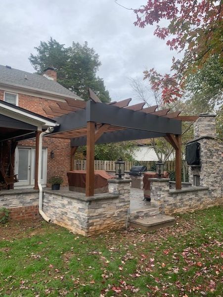 Stone patio with wooden pergola beside a brick house, with autumn leaves and overcast sky