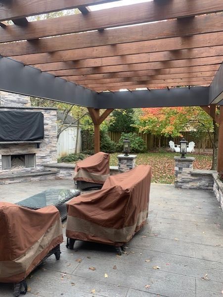 Covered patio with wicker chairs, pergola, stone fireplace, and autumn trees in a backyard.