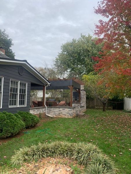 Suburban backyard with gray house, stone patio, green lawn, and autumn trees under a cloudy sky