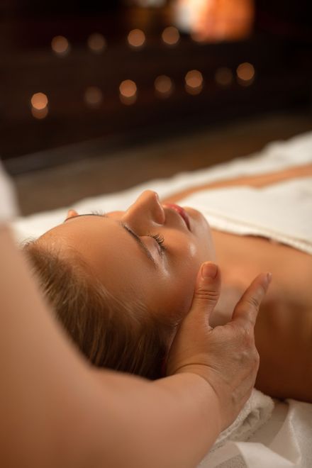 Woman receiving a facial massage at a spa. Hands on her cheek and jaw. Candles in the blurred background.