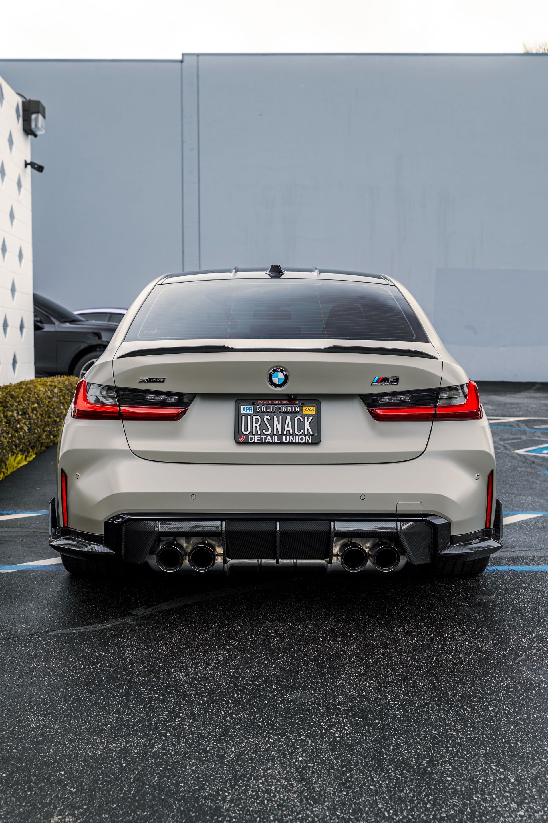 Rear view of a white BMW parked on a wet lot in front of a plain gray wall.