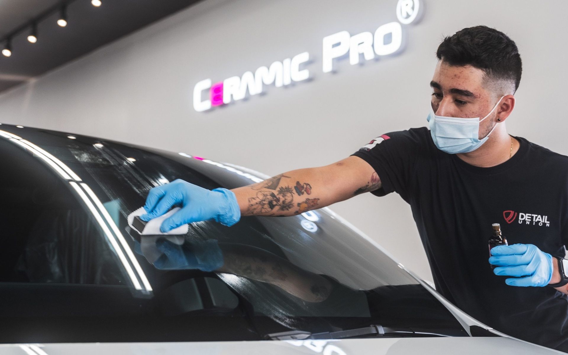 Detailing technician in a mask and blue gloves cleaning a black car windshield with a cloth.