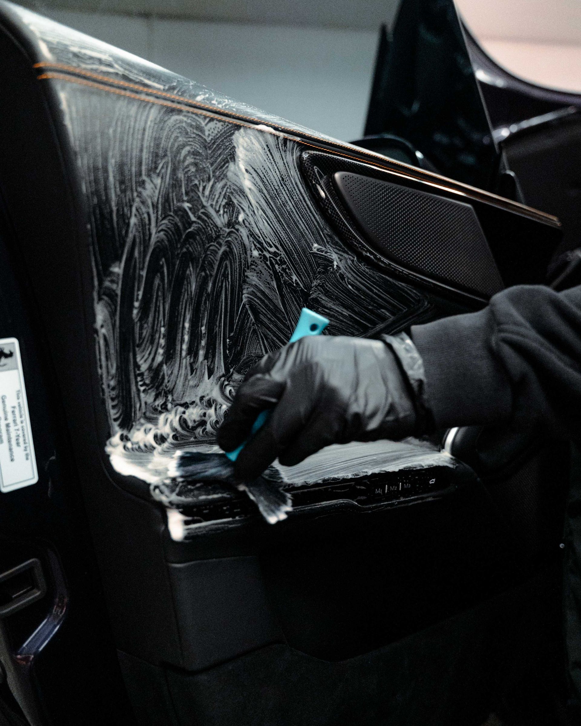Gloved hand scrubbing a black car panel with soap suds during washing