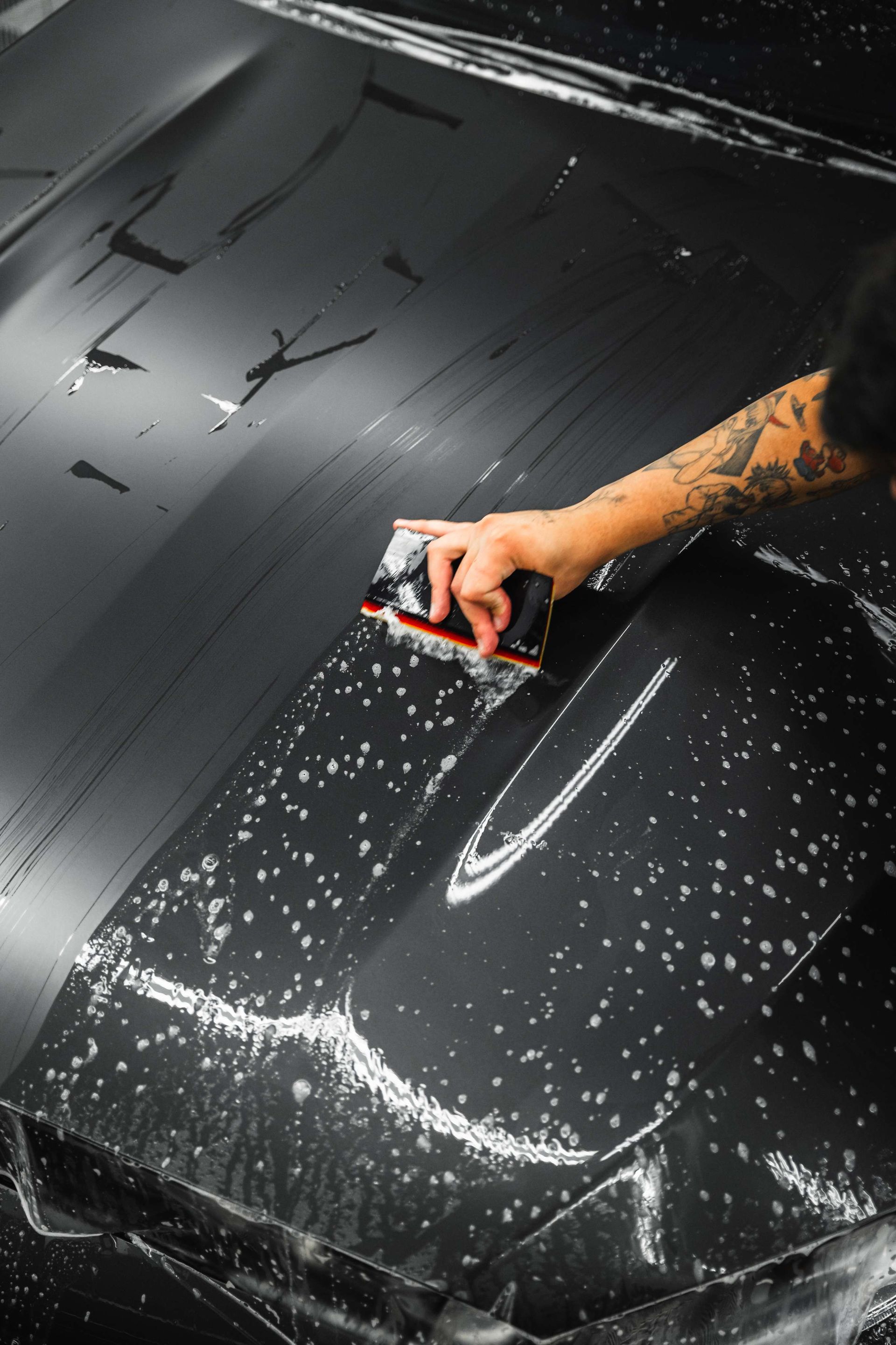 Hand smoothing black paint or vinyl on a car hood with a squeegee, leaving wet streaks and bubbles