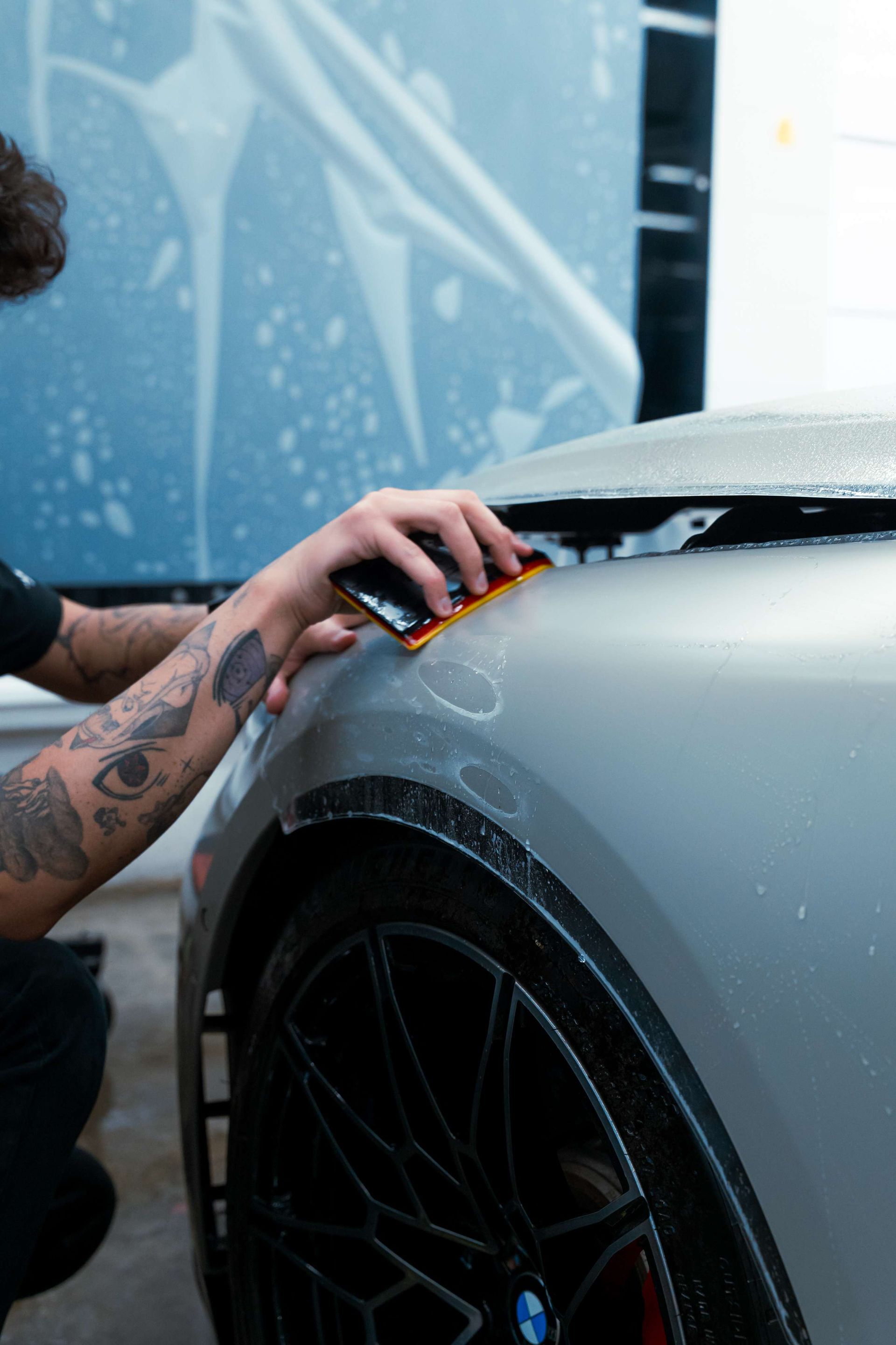 Person applying clear protective film to a blue car bumper in a workshop