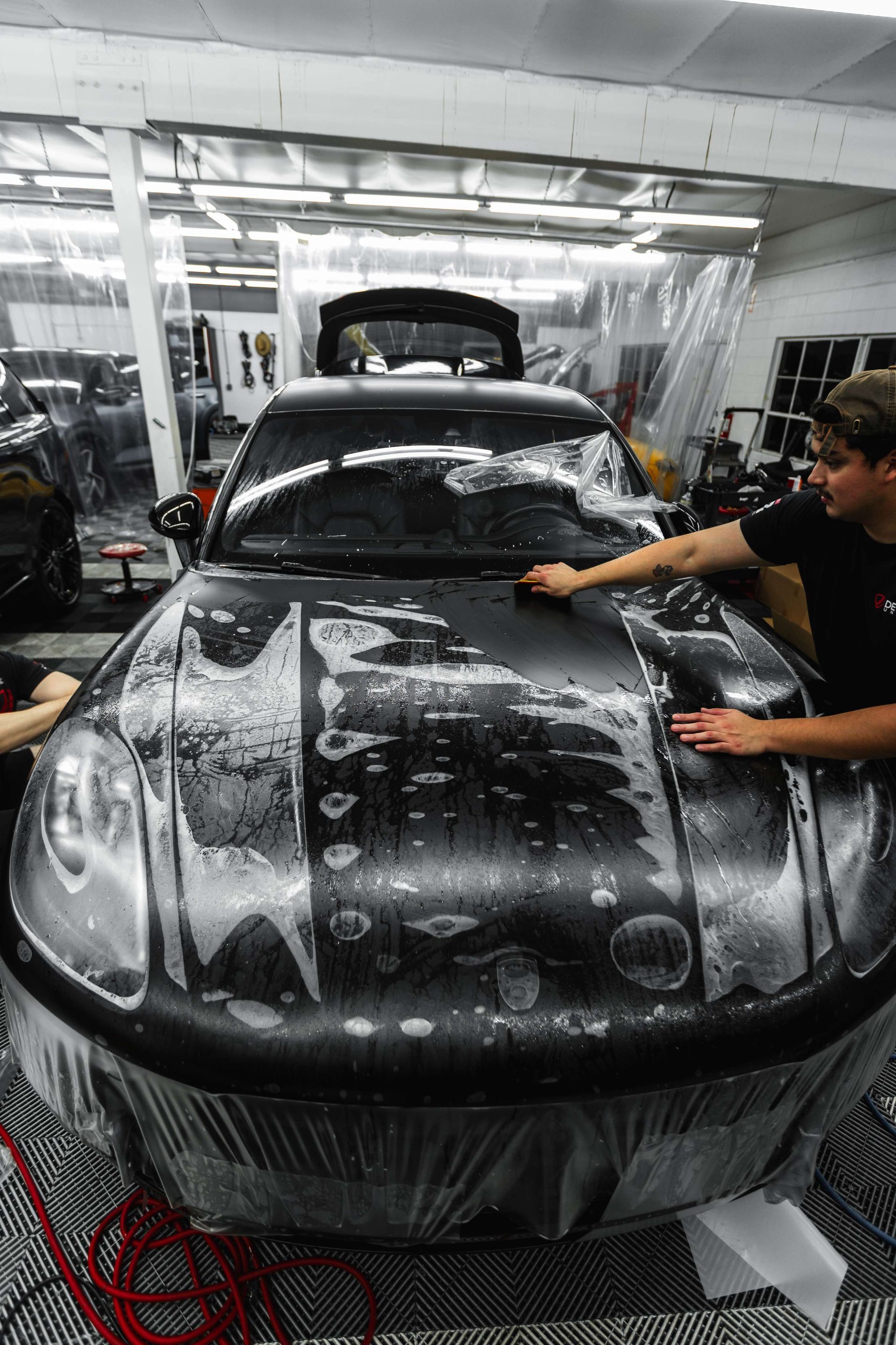 Person washing a black car in a garage with soap foam covering the hood and windshield