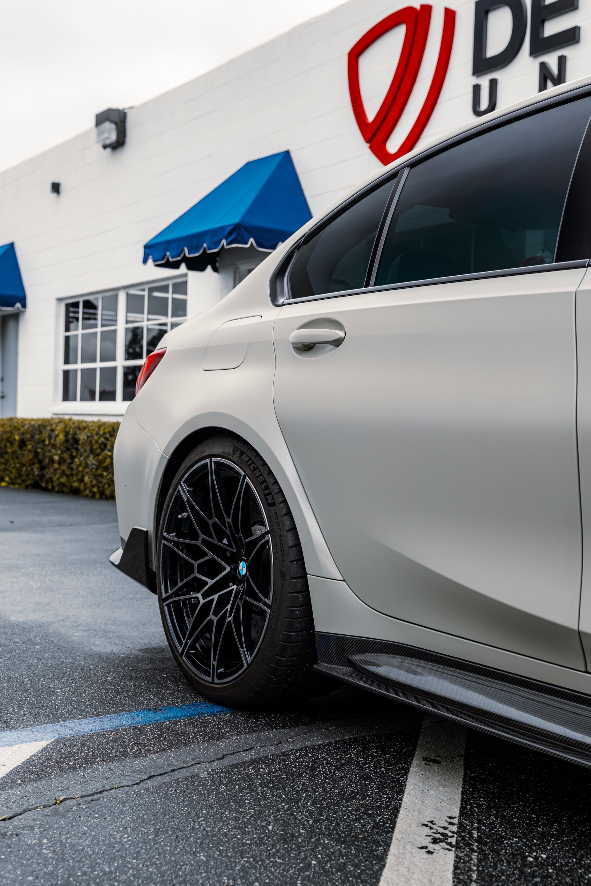 Silver sports car parked outside a dealership with a blue awning and wet pavement