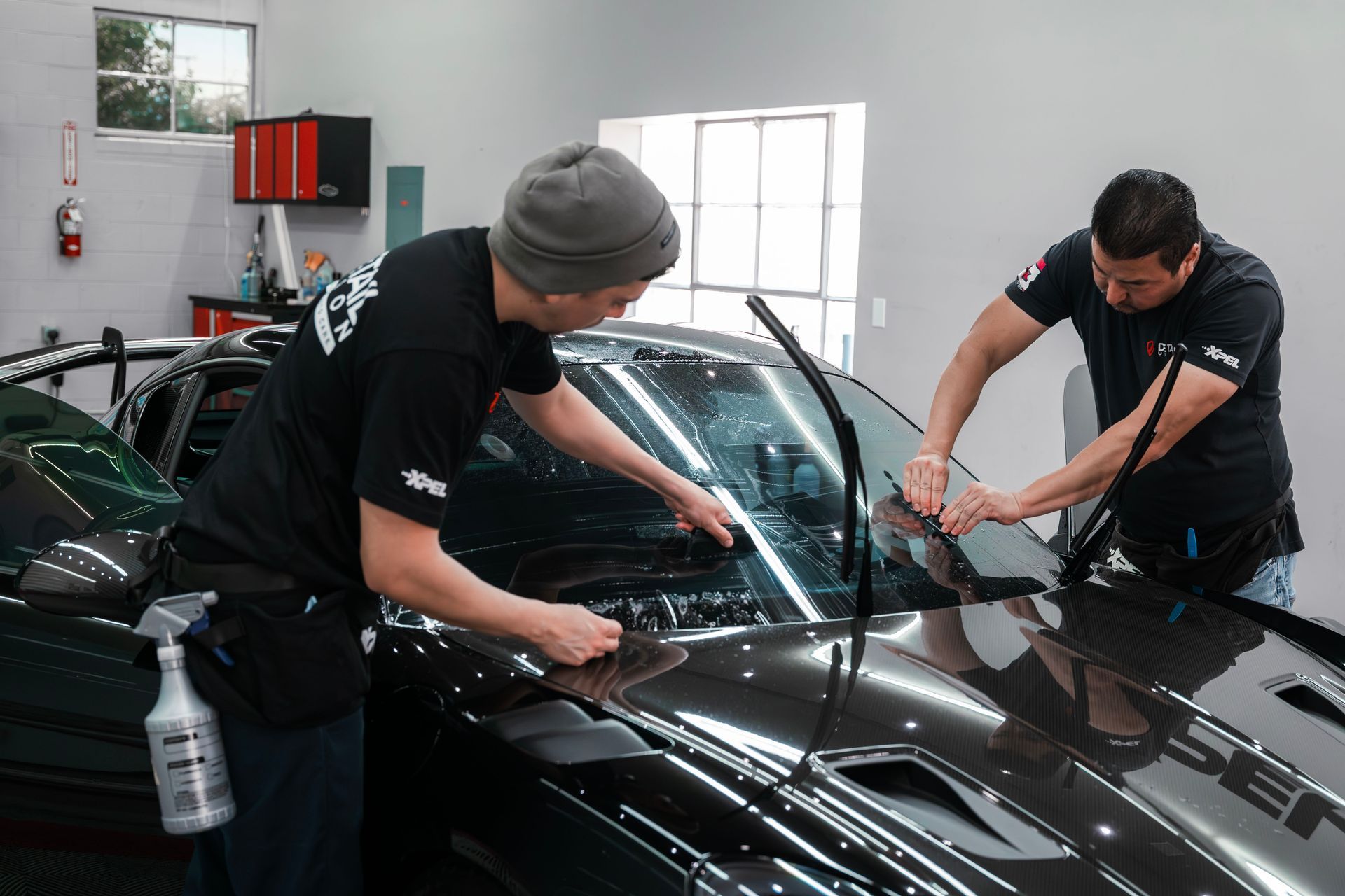 Two workers washing a black car in a garage, wiping the windshield and hood.