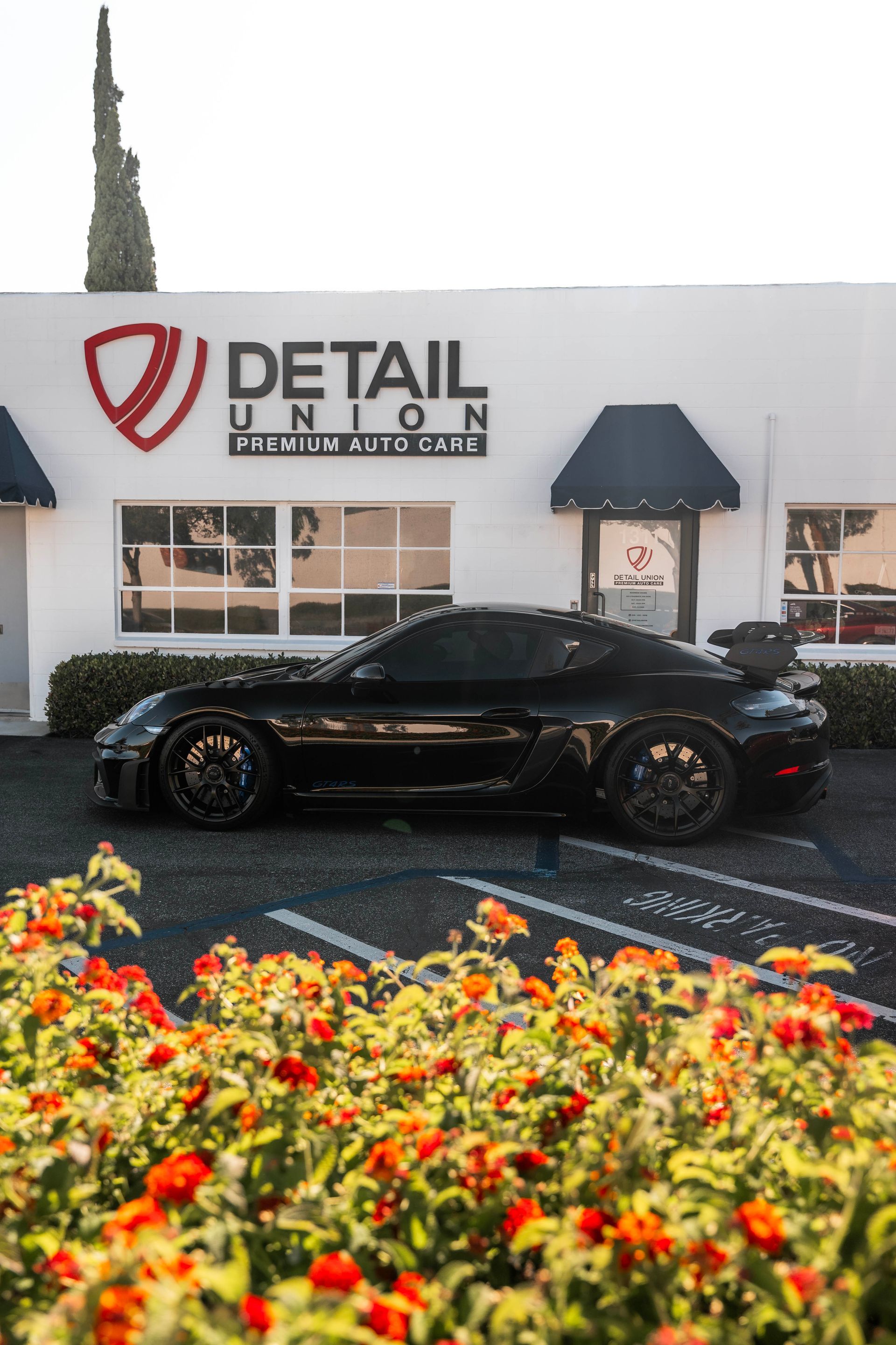 Black sports car parked outside Detail Garage, with colorful flowers in the foreground.