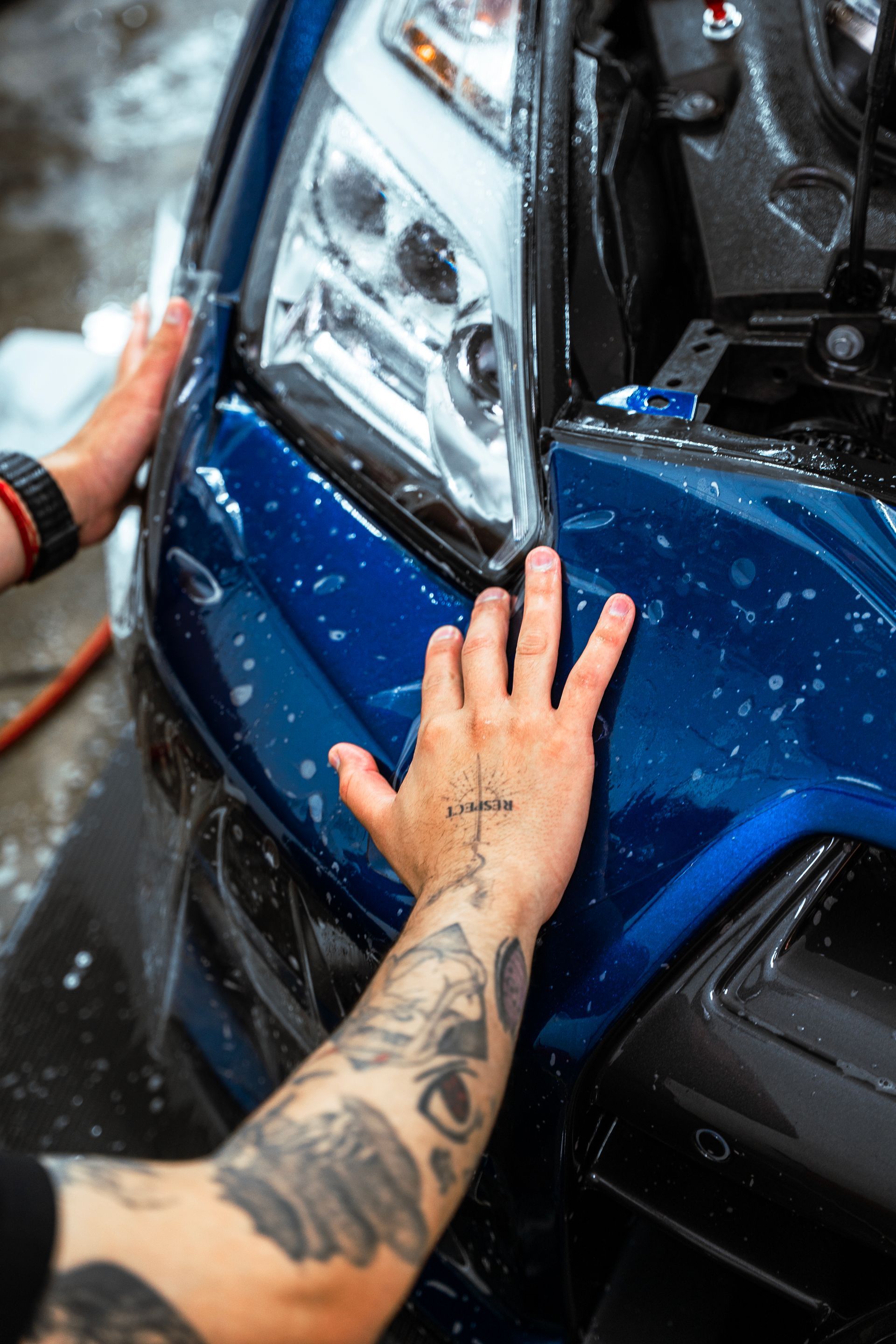 Hands washing a blue car’s front headlight and bumper with water spray