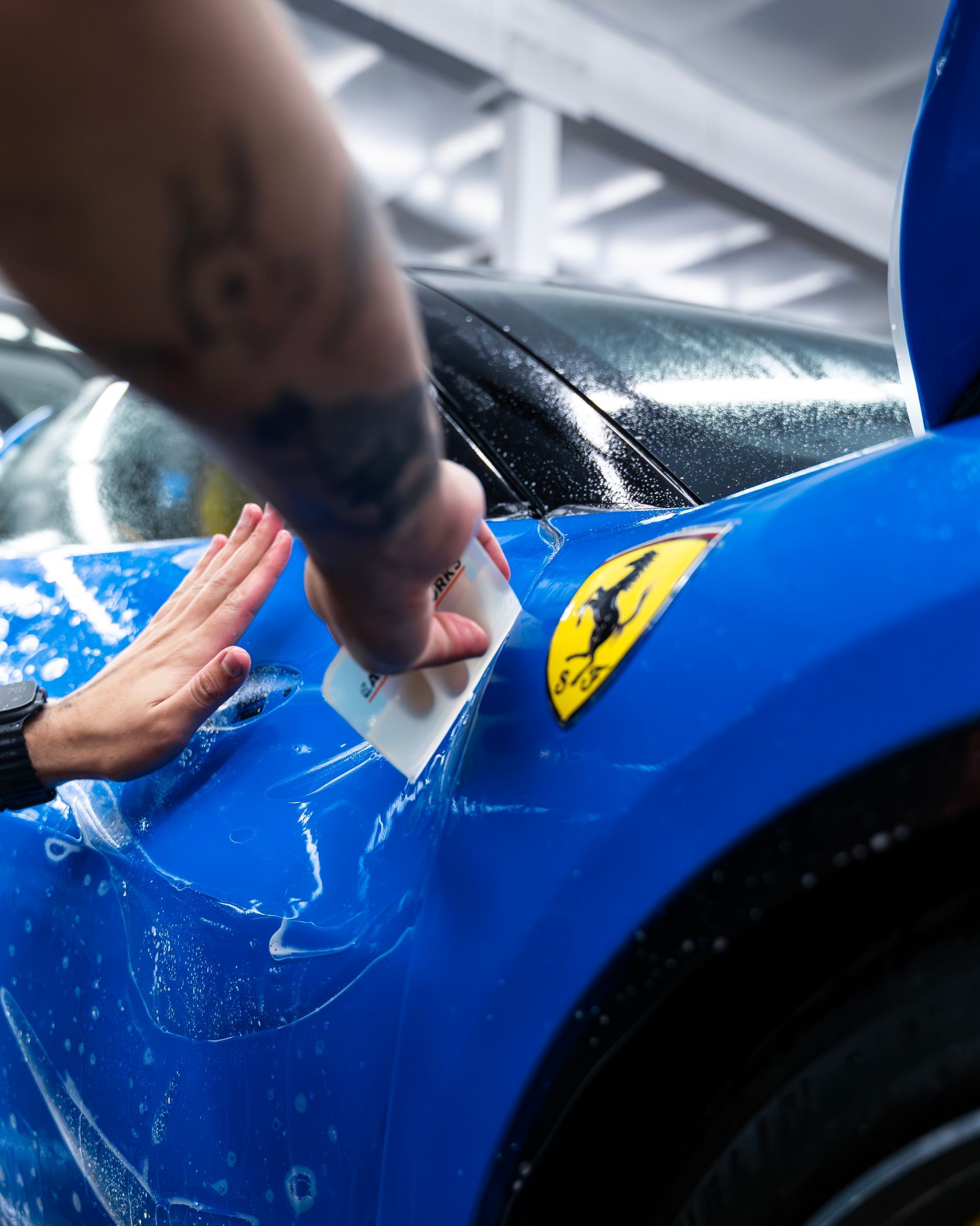 Hands applying liquid to a blue sports car’s front fender in a workshop