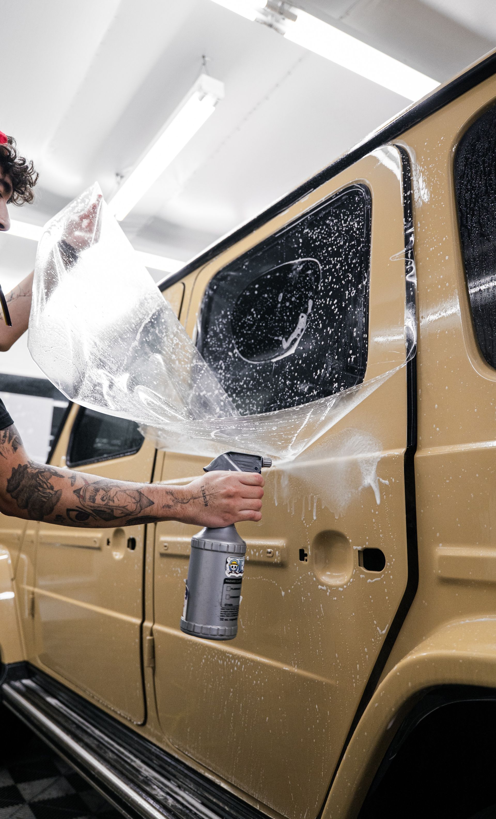 Person spray painting a gold SUV with clear protective plastic on a workshop floor