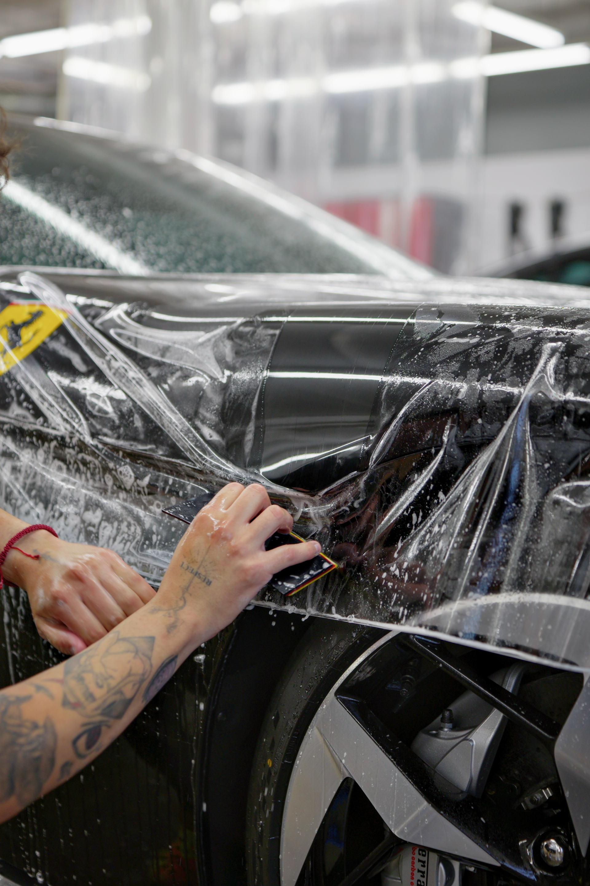 Hands applying clear protective wrap to a black car in a garage