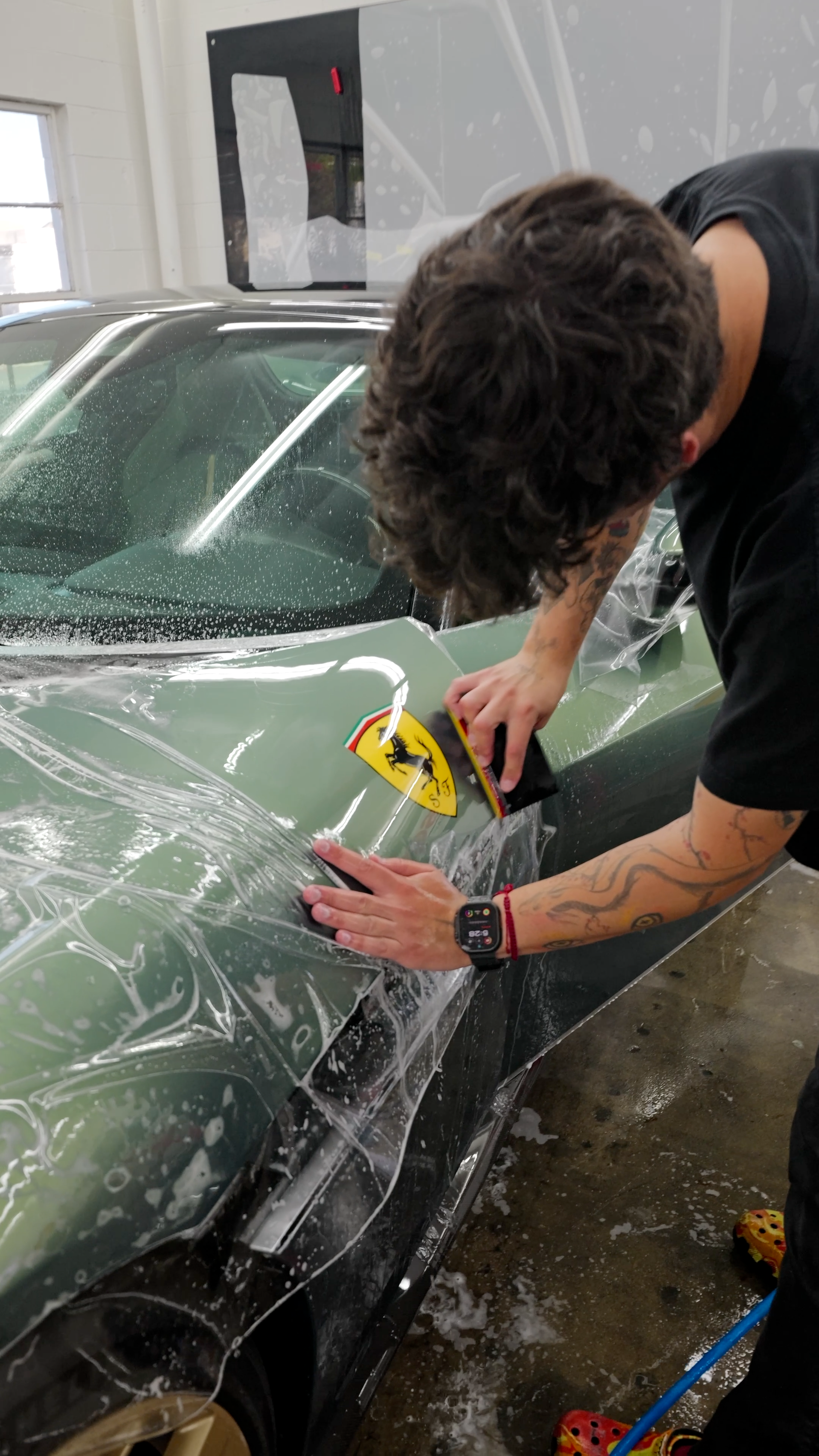 Person cleaning a dark green car windshield with a squeegee in a garage.