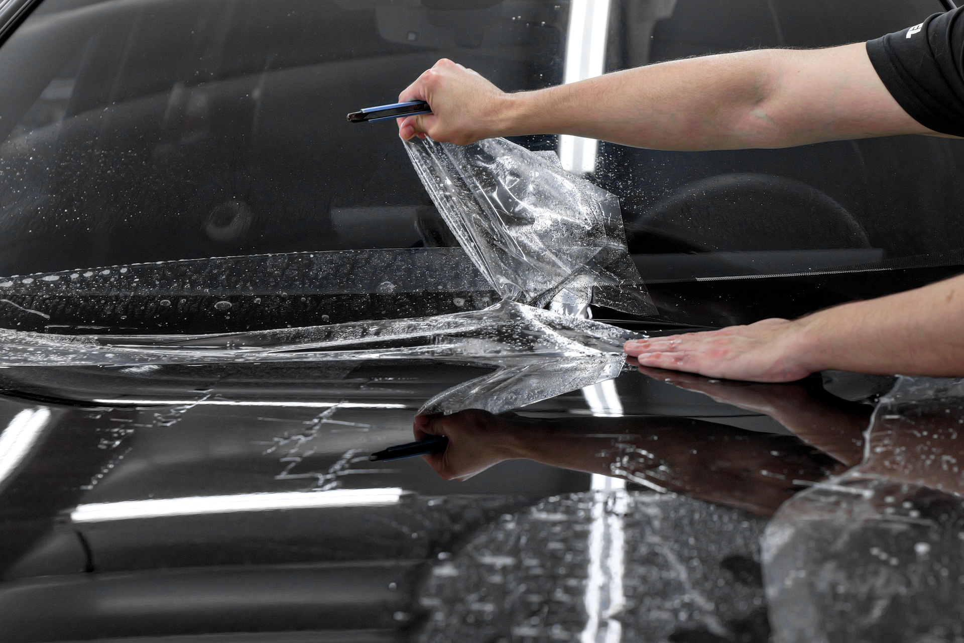 Person peeling clear protective film from a black car hood with a squeegee, water visible on the surface