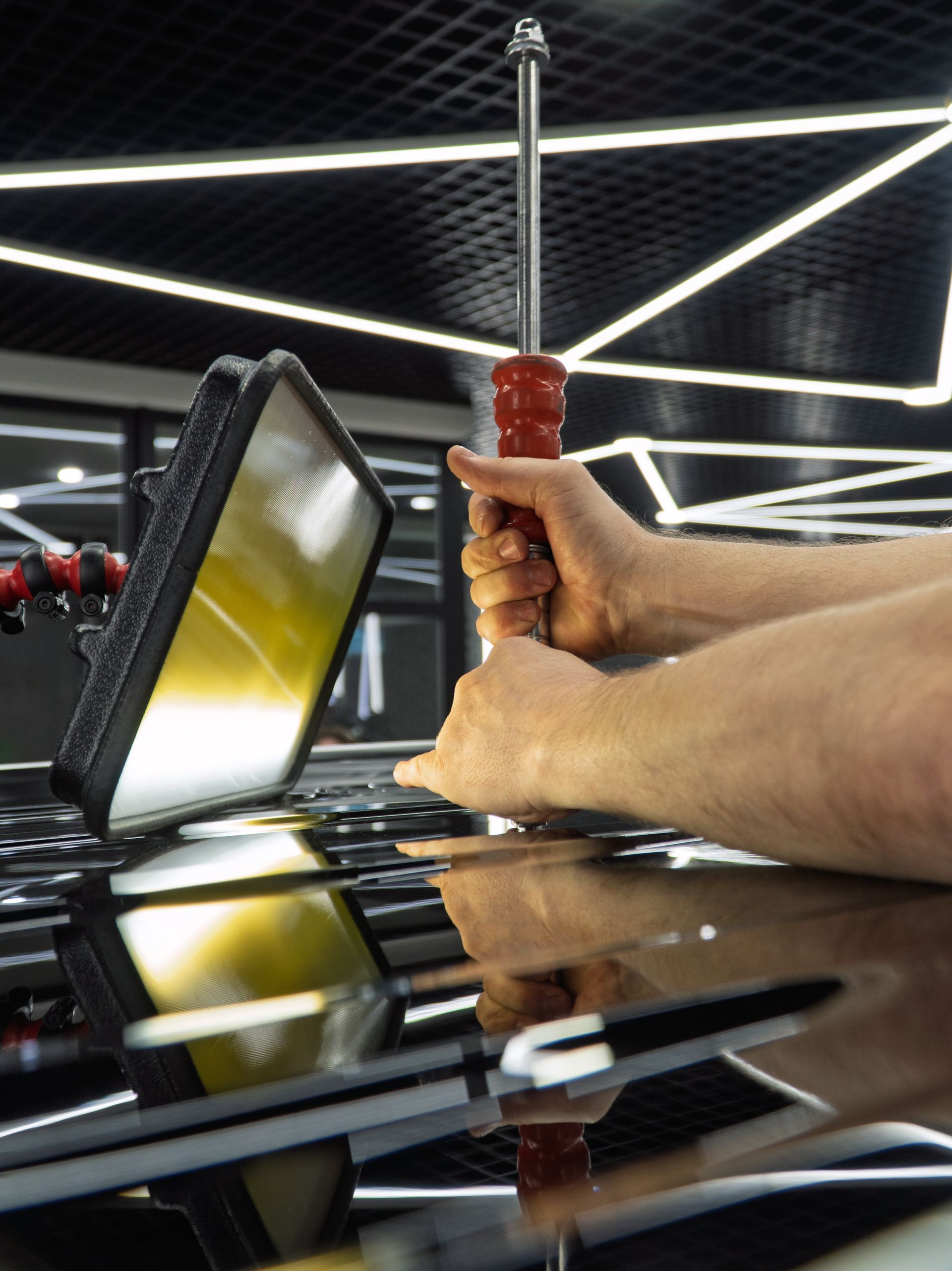 Hands polishing a black car hood with a red-handled tool under bright workshop lights