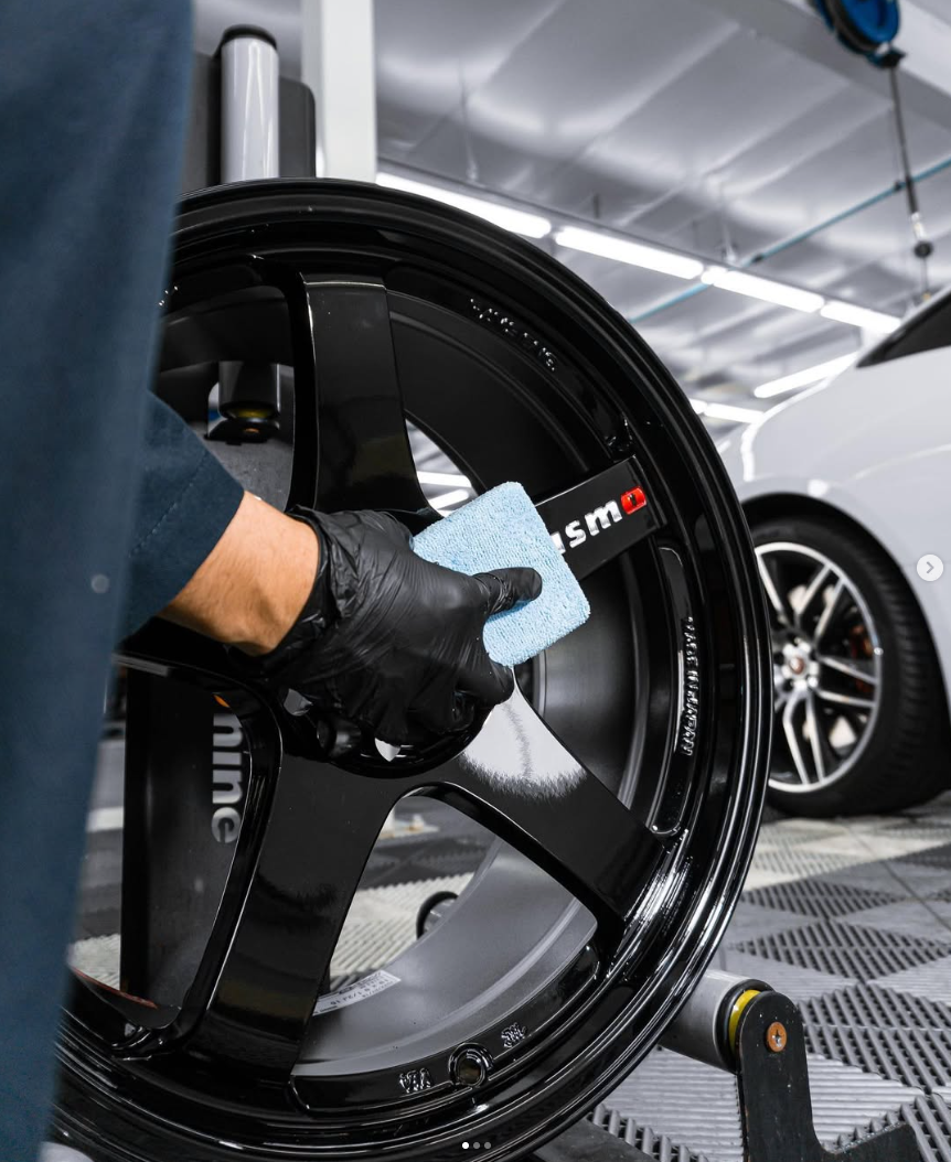 Person cleaning a glossy black car wheel with a blue cloth in a garage.