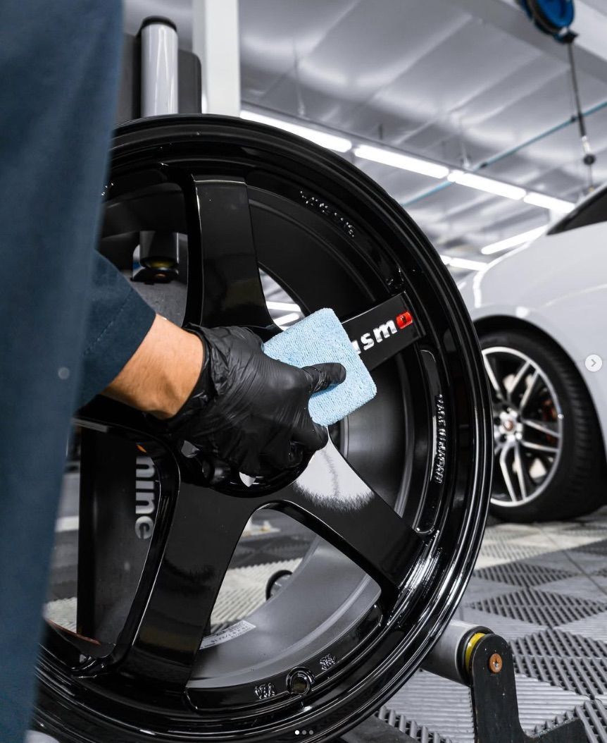 Gloved hand cleaning a glossy black car wheel in an auto detailing shop