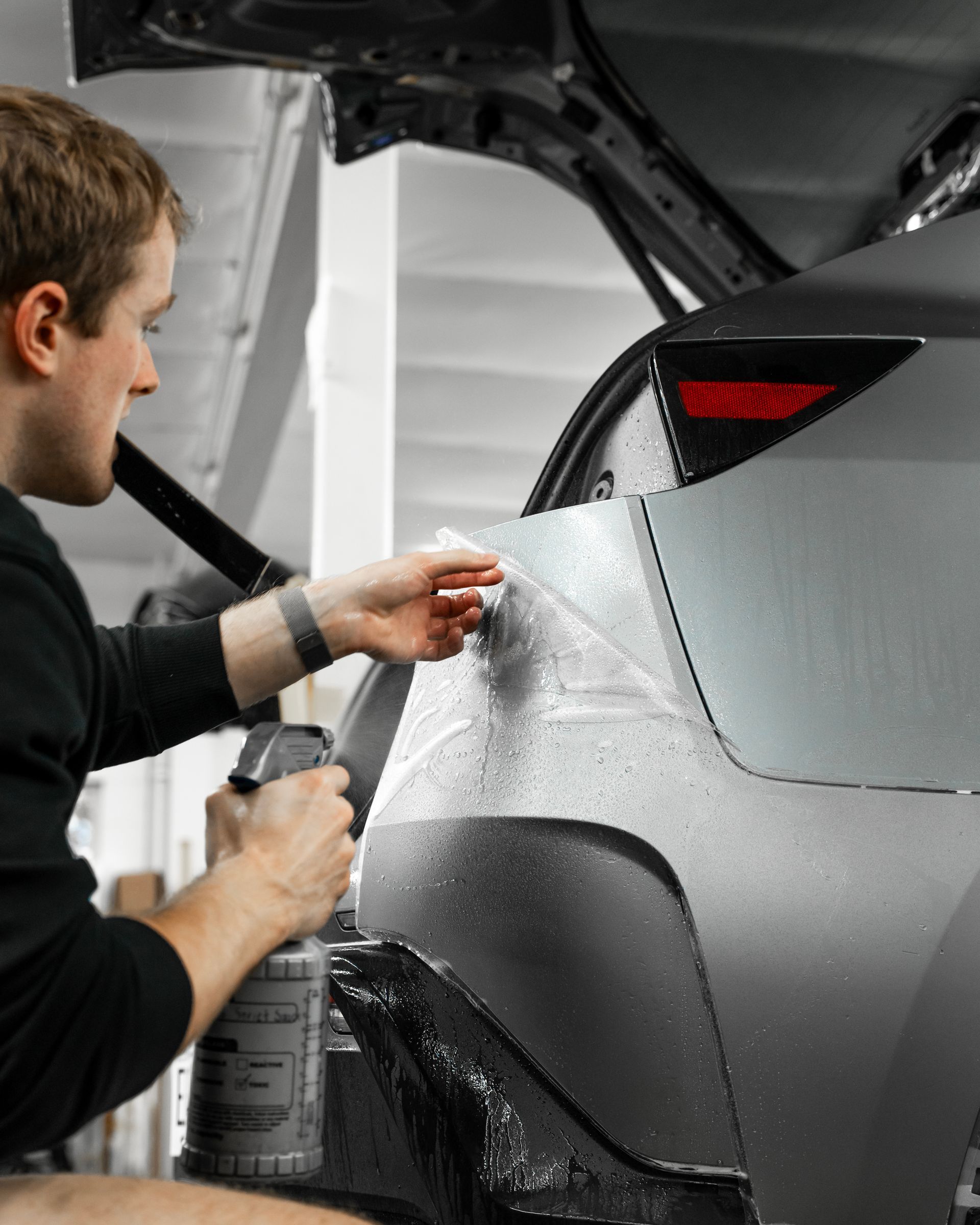 Person washing a gray car’s rear quarter panel with a spray bottle in a garage.