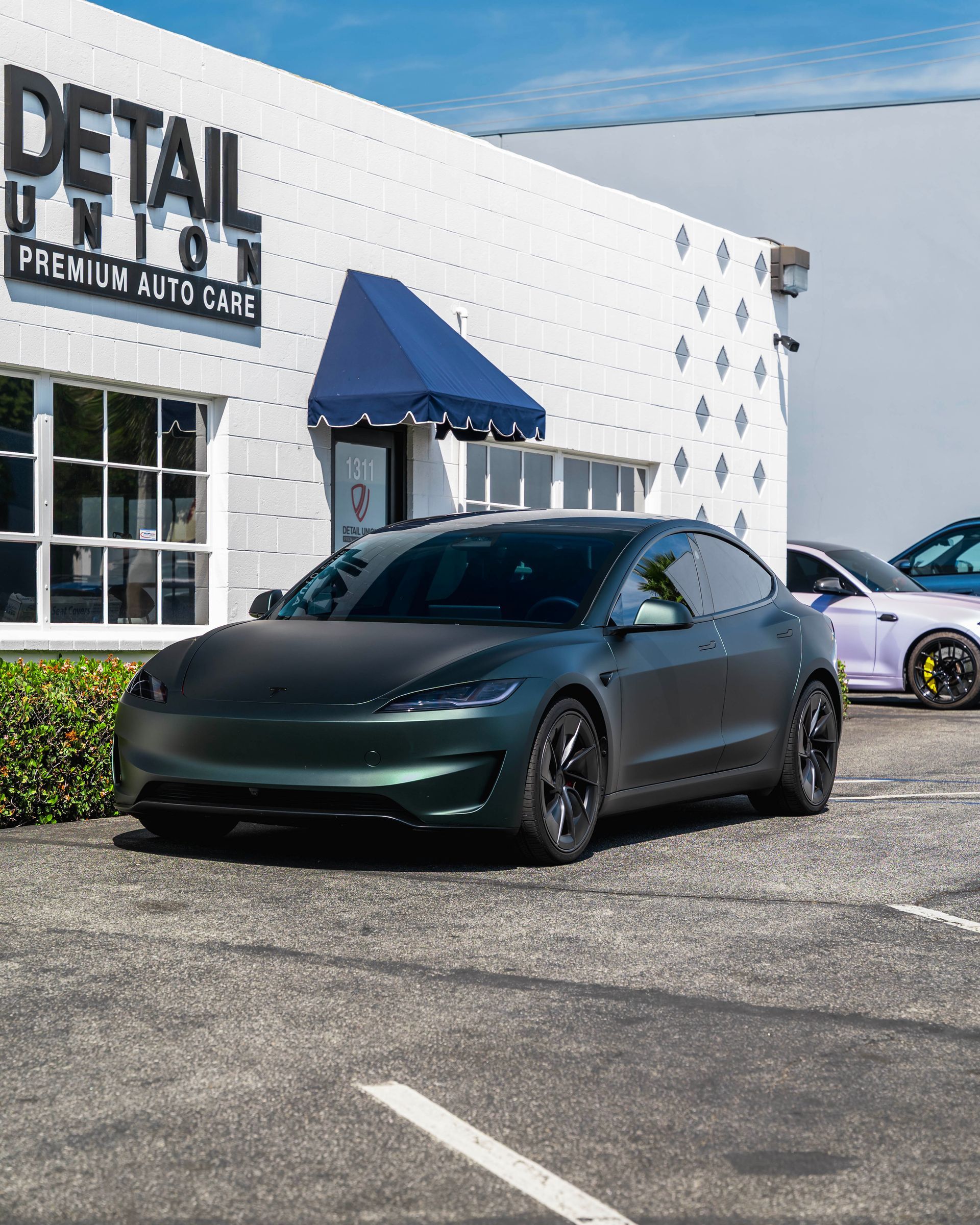 Matte black Tesla parked outside a car detail shop with a white wall and blue awning