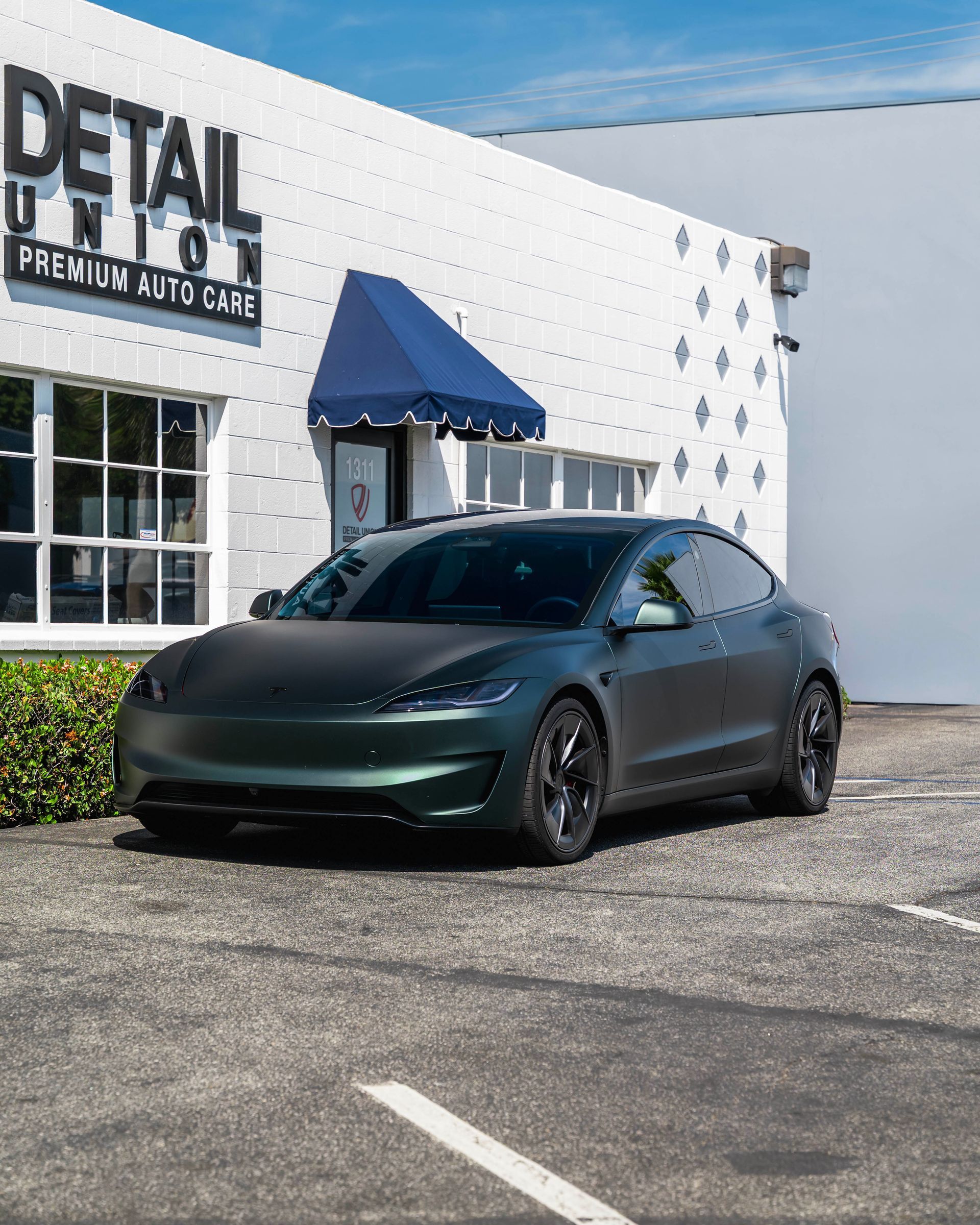 Matte dark green sedan parked outside a detail shop with a blue awning